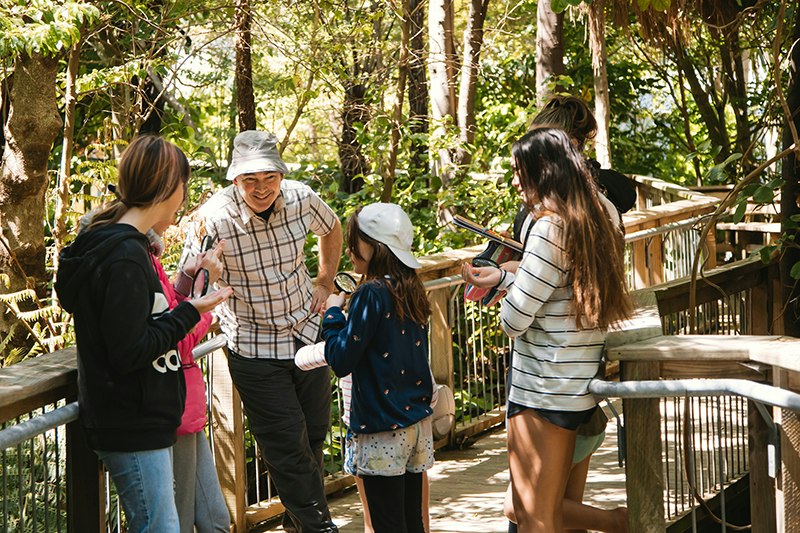 A smiling Leon in a bucket hat stands on a bridge in Bush City. He is talking to a group of 5 children holding magnifying glasses and clipboards.