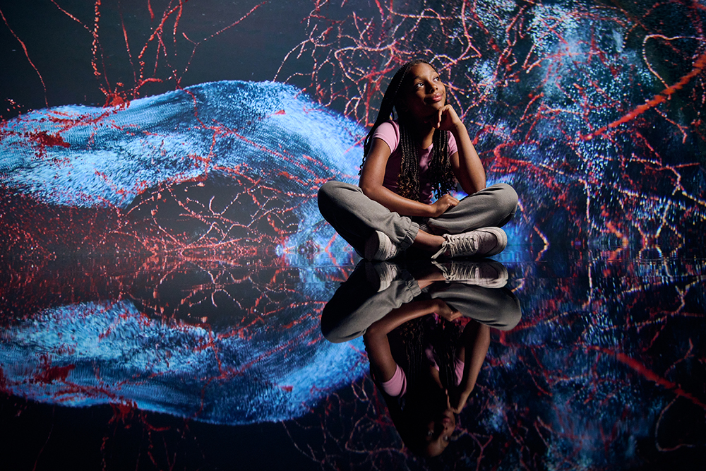 A girl is sitting cross-legged on a shiny dark surface that is reflecting the digital patterns on the wall behind her.