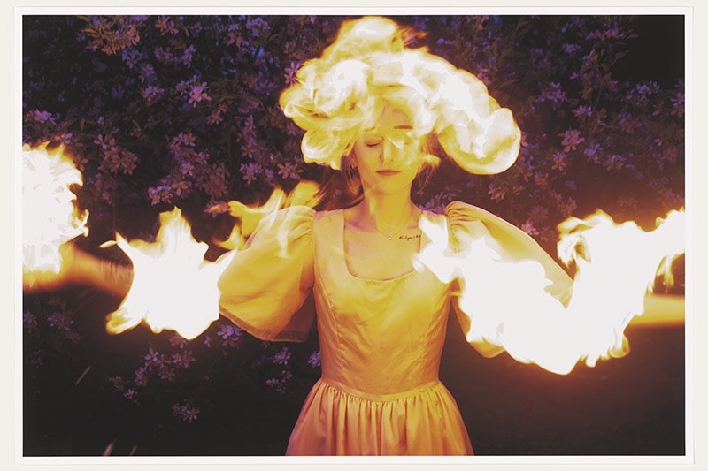 A woman wearing a silk gown with puffy sleeves stands with arms outstretched in front of a background of purple flowers. Flames engulf her head and arms while she smiles serenely with eyes closed.