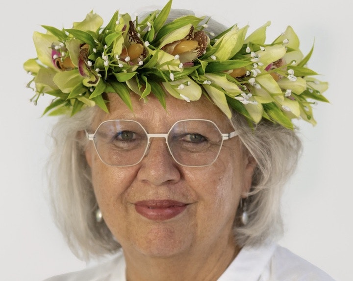 A headshot of a woman with shoulder-length grey hair and glasses on. She has an 'ei, or floral wreath on her head.