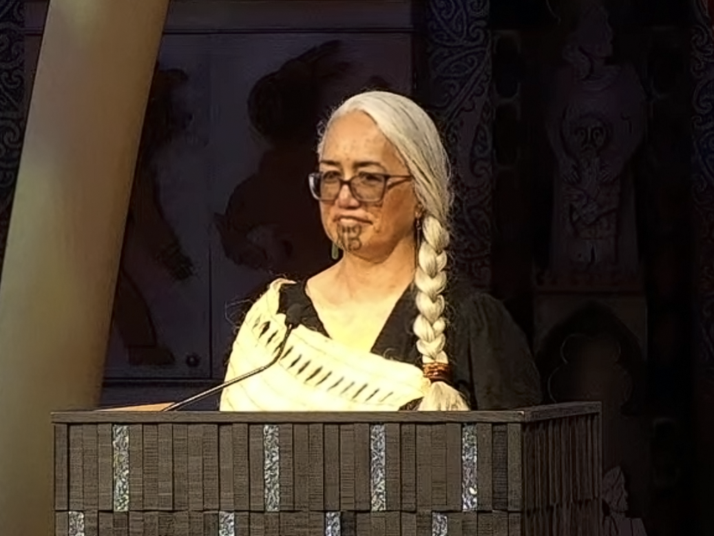 Still of Dr Awhina Tamarapa. Te Papa The head and shoulders of a woman standing at a podium. She has a cloak draped over one shoulder, and her hair is in a thick plait on her left side.