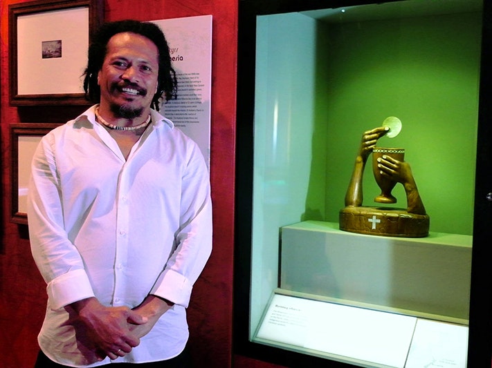 Selwyn Teho and 'The Great Elevation' chalice and host. Photo by Rowan Moulder. Te Papa A dark skinned Solomon Island man in a white dress shirt stands beside a carved sculpture. The sculpture is dark timber chalice and 2 arms, inlaid with shell. One arm holds the chalice and the other holds a communion wafer made of shell.