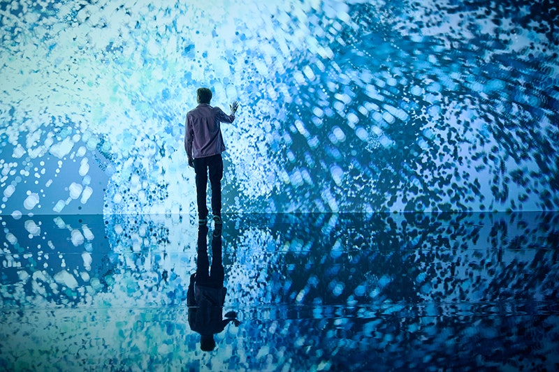 Man standing in front of immersive installation of swirling blue light witha  hand raised to touch it.