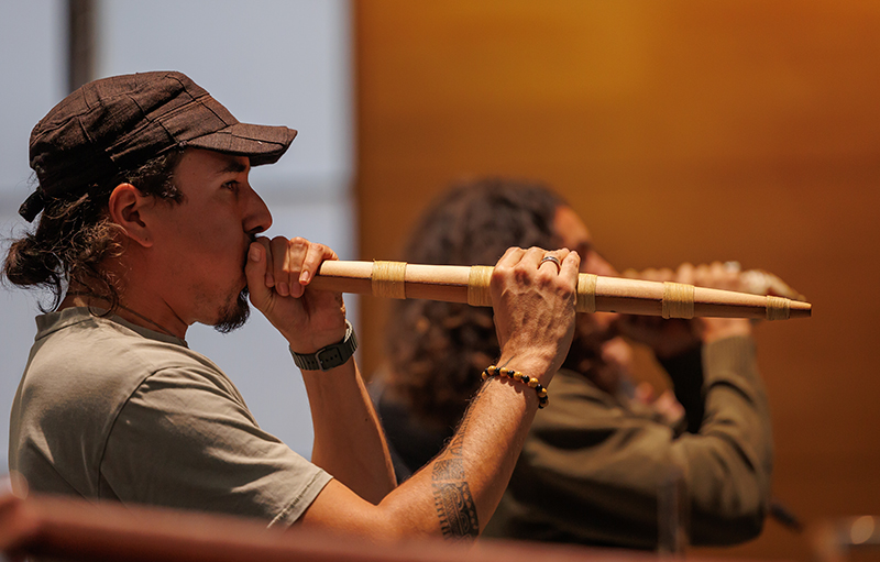 A man with long hair, a beard and cap is in the foreground playing a pūkāea - a long, narrow traditional Māori taonga puoro instrument, used to welcome people. A second man plays a taonga puoro instrument behind him.