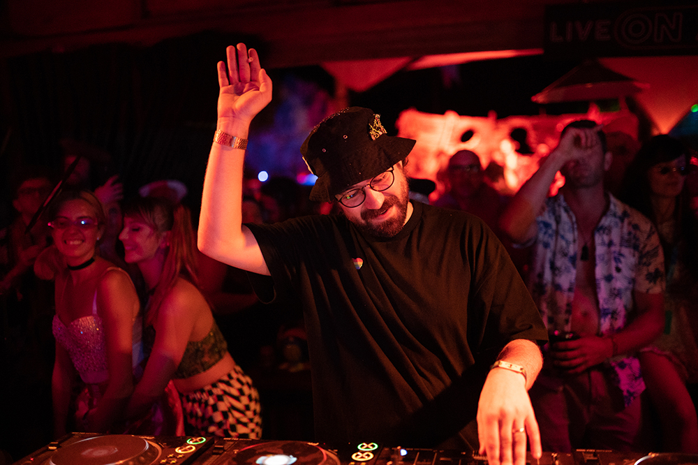 DJ raises one hand while mixing on the decks, playing to a tightly packed crowd dancing under warm red lighting.
