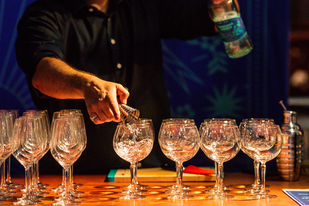 Bartender pours liquid from a small cocktail measuring tool into a row of large stemmed glasses.