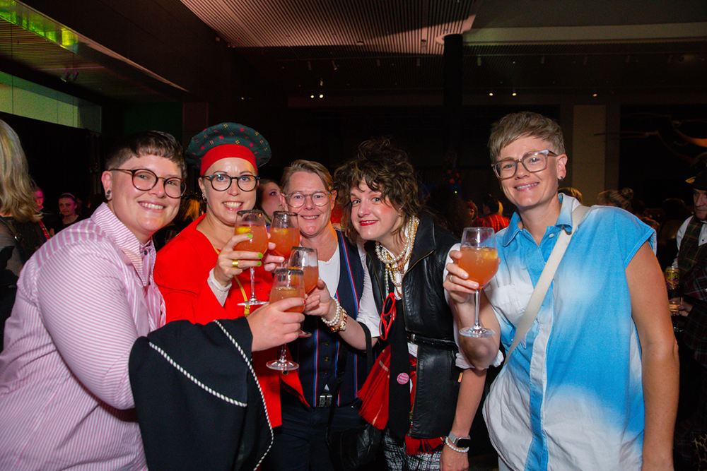 Five people smile and raise cocktail glasses in a toast at a dimly lit indoor event. They wear glasses and eclectic clothing including tartan, leather, and bold colours.