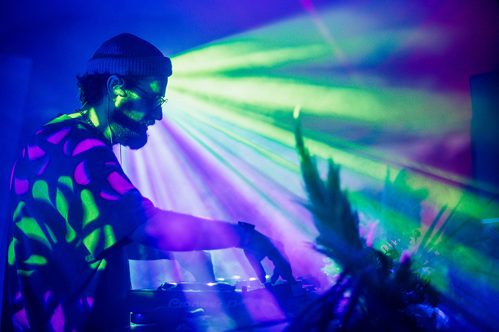 DJ adjusts the mixer under sharp green and purple lights, with beams cutting through haze and patterned light shifting across his shirt.