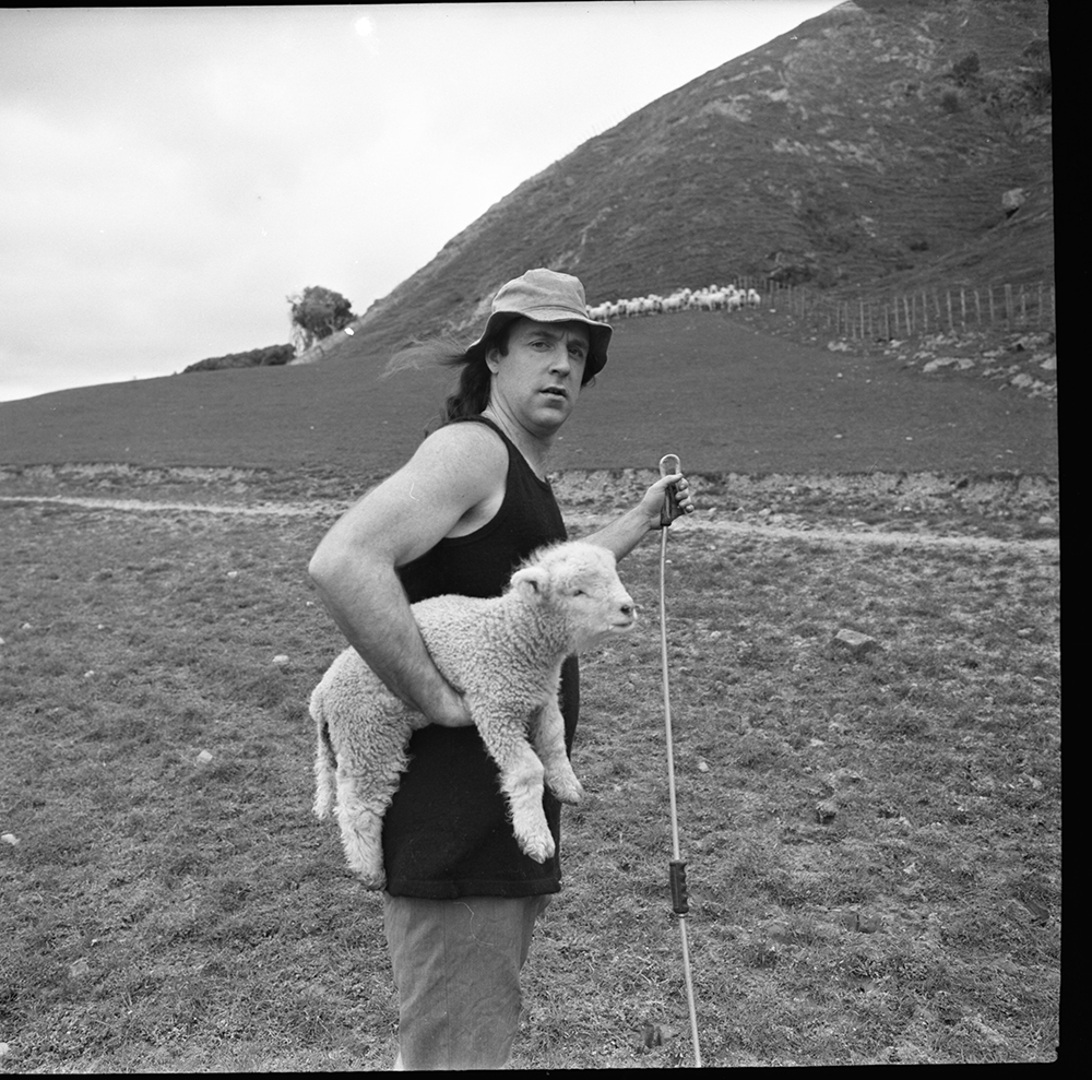 A black and white photo of John Clarke as Fred Dagg on a farm carrying a lamb under his right arm.