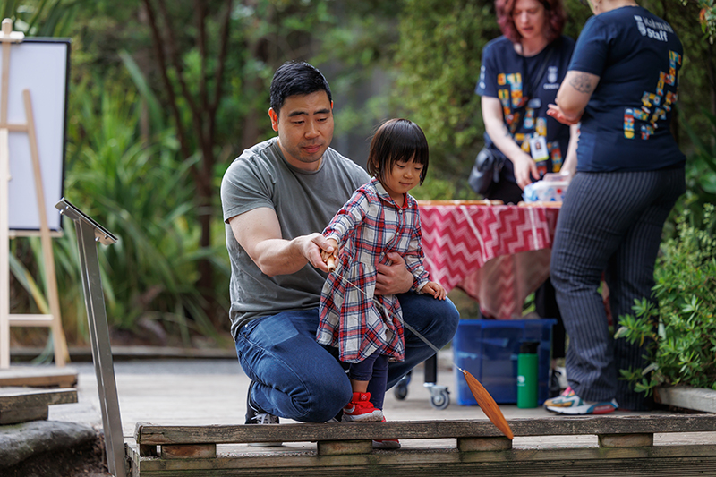 Man with little girl playing and whirling a wooden instrument outside.