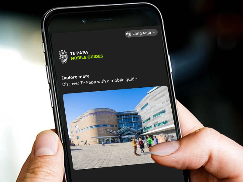 A close up of a hand holding a mobile phone that has an image of Te Papa from the front forecourt.