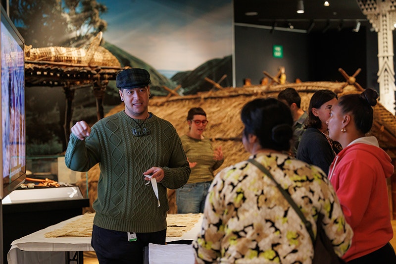 A male Curator is standing in an exhibition facing the camera, wearing a flat cap, green sweatshirt and a hei tiki pounamu taonga around his neck. He speaks to two women who have their back to the camera, holding a small piece of material in his hand. 