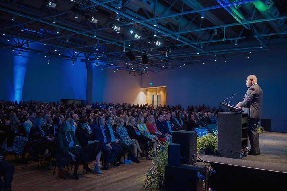 A large auditorium with a man in a suit standing at a podium talking to a large seated audience.