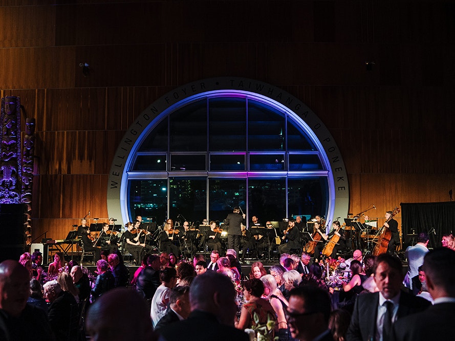 Spring Soiree, 2024. Photo by Smoke Photography. Te Papa (247827) A large room with a round window is full of people at a party. The room is light in dark purples and blues.