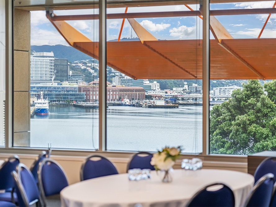 Te Papa Venues, 2024. Photo by The Way Home. Te Papa (273343) A white-clothed table with blue chairs around it next to a window with a view of Wellington Harbour.