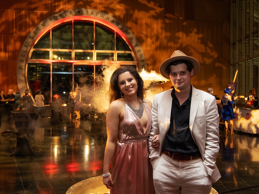 Surrealist Soiree, 2021. Photo by Jo Moore. Te Papa (175838) Two people are standing together in the foreground and smiling at the camera. There is an event with dry ice in going on in the background.