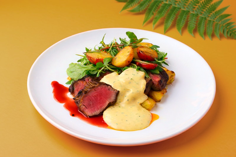 A plate on an orange table with sirloin steak, green salad, potatoes and a creamy sauce. There is a green fern in the top right-hand corner of the table.