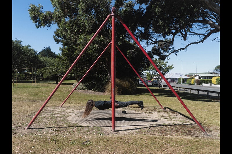 A person with long hair is lying flat on a swing in a park. It looks like they are hovering above the ground.