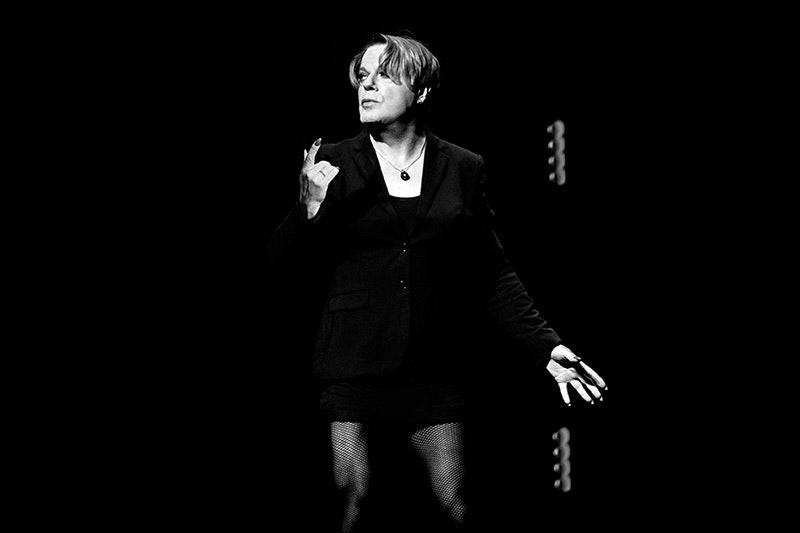 A person in fishnet stockings and a suit jacket is on a dark stage under a spotlight.