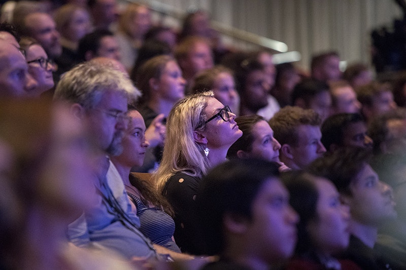 Audience members sit closely packed in tiered seating, watching a speaker off camera under low auditorium lighting.