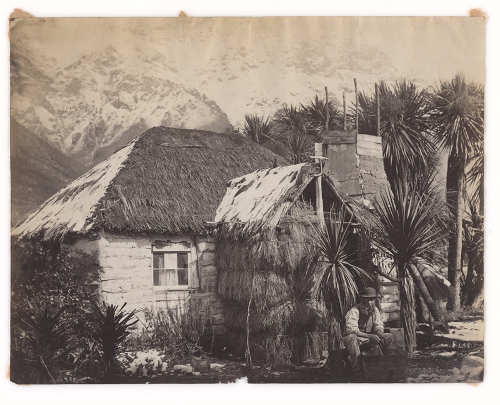 A sepia photograph of a cottage with cabbage trees outside and a man sitting out the front.