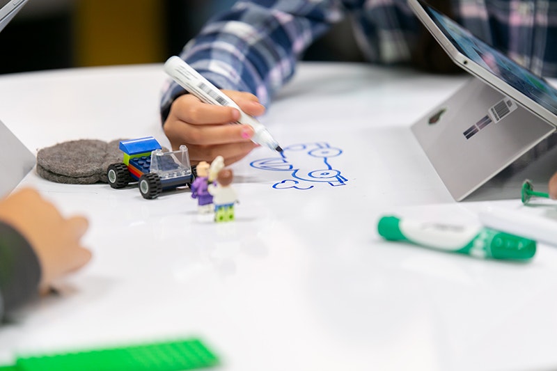 An image of a child's hand drawing with a marker pen. There is a tablet next to him.