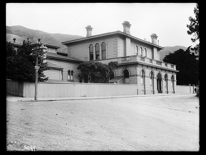 Colonial Museum; 1904, Augustus Hamilton. Te Papa (MA_C.001054;) A black and white photo of a wooden building with three chimneys viewable, and a wide street in front of it.