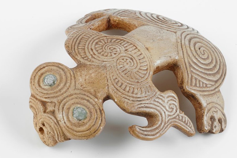 A small carving of a stylised figure with paua eyes sitting on a white surface.