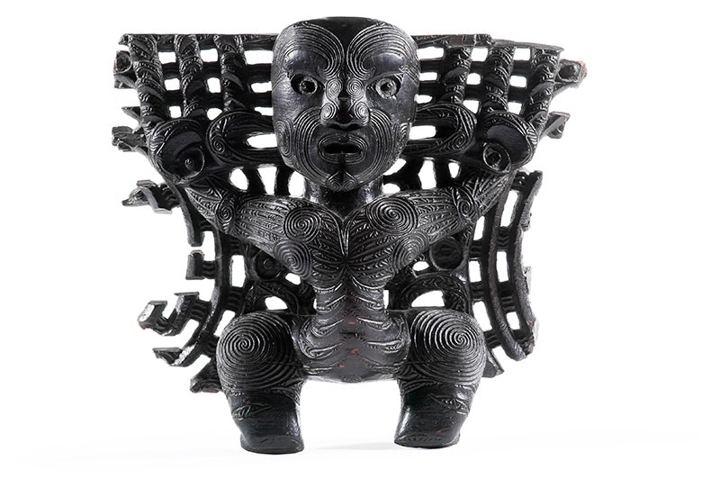A dark brown or black piece of Māori wood carving on a white surface.