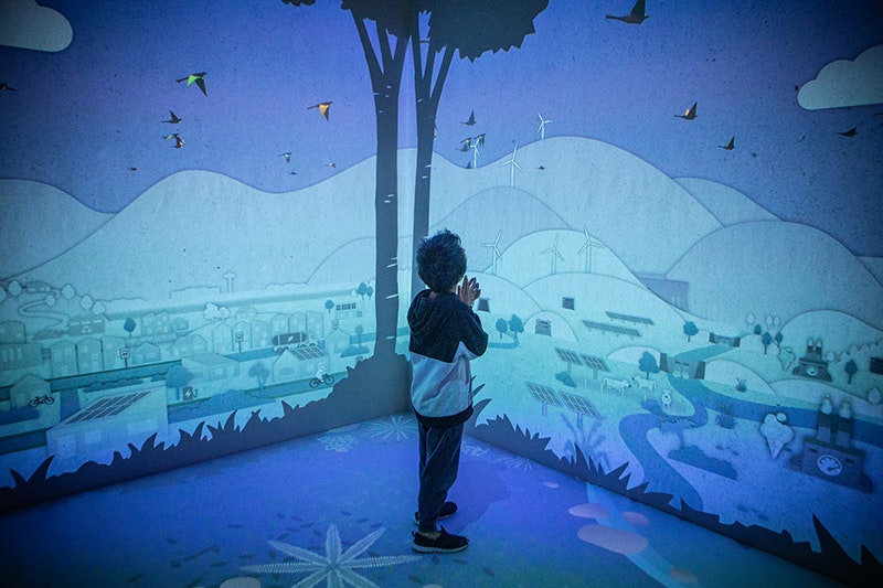 A chid is standing in a room with a full projection of turbines and trees.