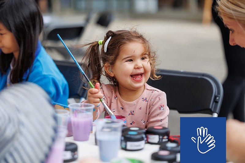 A close-up image of little girl, aged around three years old, holding a paint brush while smiling broadly at a lady to her right.
