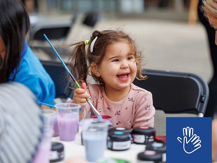 Raumati Whānau Day 2025, Photo by Jo Moore. Te Papa (257102) A close-up image of little girl, aged around three years old, holding a paint brush while smiling broadly at a lady to her right.