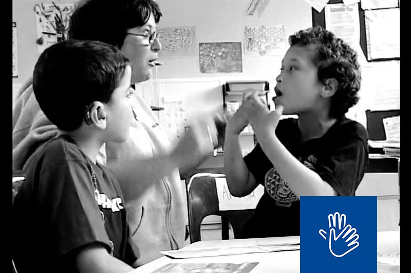 A black and white image of a teacher signing to a deaf child who is also signing.