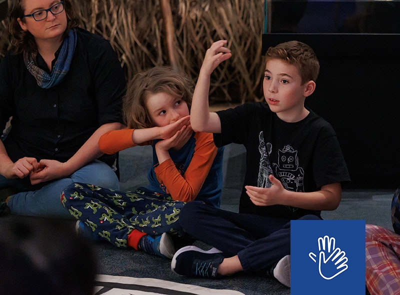 A child is sitting on the floor and making actions with his hands. There is another child next to him and an adult.