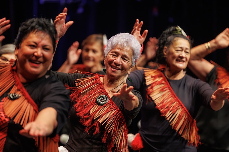 Tūhourangi Ngāti Wāhiao Koeke, Taikura Kapa Haka 2025. Photo by Jo Moore. Te Papa (270891) Three women in front of other women performing a song with hand actions. They all have the same red sash on.