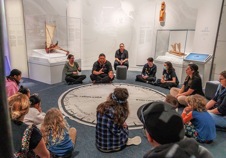 Several people sitting around a circular mat showing ways to navigate. There is one person speaking in English, and one person using NZSL.
