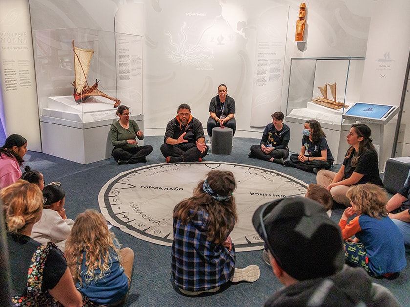 Matariki Whānau Day 2025. Photo by Jo Moore. Te Papa (268360) Several people sitting around a circular mat showing ways to navigate. There is one person speaking in English, and one person using NZSL.