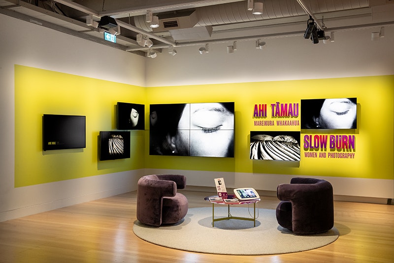 2 low, cushioned, crushed velvet armchairs with rounded half backs next a small touch screen on a low round table atop a round rug on hardwood floor. 5 bright screens are mounted on the back corner walls. The exhibition name Ahi Tāmau | Slow Burn is in uppercase purple and red letters coloured against a chartreuse rectangle. The screens and seating are under spotlights causing strong shadows.