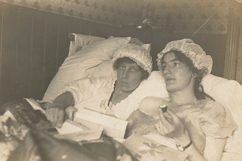 A sepia photograph of two gender non-conforming people lying side by side in bed wearing night gowns and lace mob caps with the covers pulled up to their stomachs, pillows piled high behind them. Daisy Tinney, on the left, stares directly at the viewer, right hand resting on the covers, a ring prominent on her index finger. On the right, Daisy's friend looks ahead, nightgown slipping off one shoulder. She holds a book open in one hand and a half eaten treat in the other. Wrapping paper is strewn over the comforter.