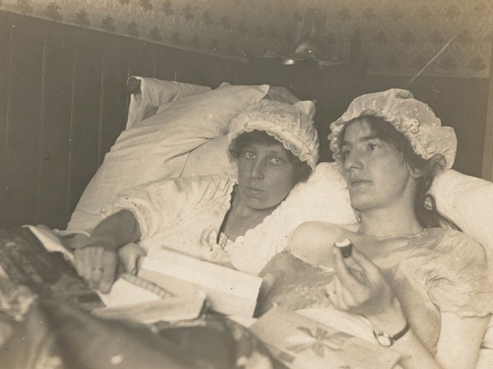 Daisy Tinney, In bed - self-portrait with friend, 1910s-20s, Wellington. Purchased 2023. Te Papa (PS.004025) A sepia photograph of two gender non-conforming people lying side by side in bed wearing night gowns and lace mob caps with the covers pulled up to their stomachs, pillows piled high behind them. Daisy Tinney, on the left, stares directly at the viewer, right hand resting on the covers, a ring prominent on her index finger. On the right, Daisy's friend looks ahead, nightgown slipping off one shoulder. She holds a book open in one hand and a half eaten treat in the other. Wrapping paper is strewn over the comforter.