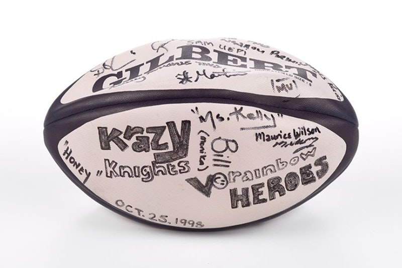 Ink signatures on a black and white, vinyl, 'Gilbert' brand rugby ball. Large block letters read "Krazy Knights v Rainbow Heroes" alongside a smiley face and the date  "Oct.25.1998". Visible signatures include "Honey", "Ms. Kelly", "Bill (Monika)", Maurice Wilson" and " Mu".