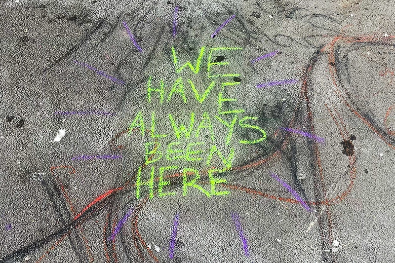 Lime green chalk writing on pavement. Text reads We have always been here. in uppercase block letters