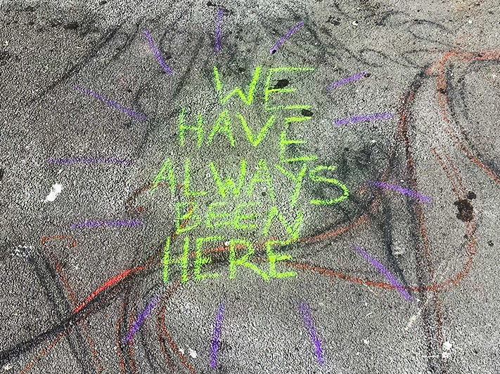 We have always been here, 2025. Photo by Will Hansen. Lime green chalk writing on pavement. Text reads We have always been here. in uppercase block letters