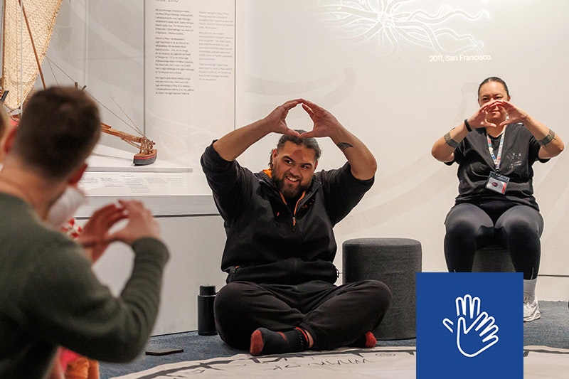 Two people are facing an audience sitting on the floor and holding their hands up in a circle shape.