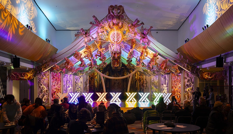 A room with coloured lighting and colourful carving behind the stage. On the stage are nine lights in x shapes.