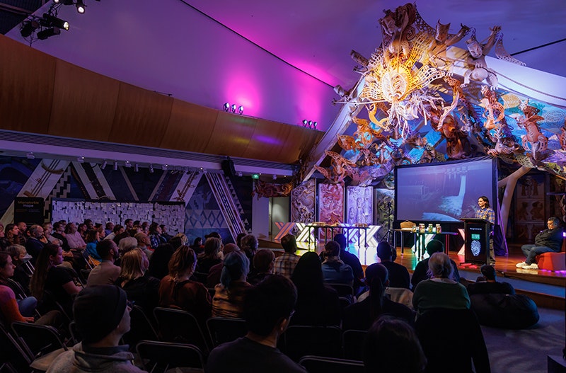 A person is standing on a colourful stage with a display screen behind him. He is talking to an audience.