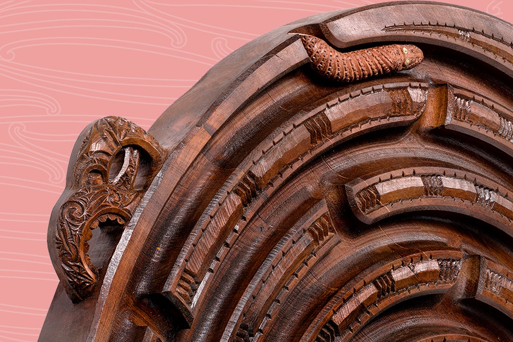 A carved wooden spiral with Māori designs on a dusky pink background.