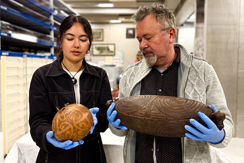 Two people wearing blue gloves are both standing in a museum store room holding carved treasures.