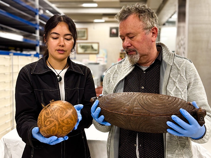 He Toi Whakairo taonga. Photo by Tia Nepia-Su'a. Te Papa (278801) Two people wearing blue gloves are both standing in a museum store room holding carved treasures.