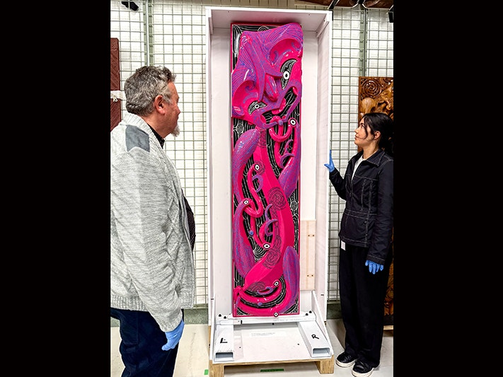 Exhibition co-curators Dougal Austin and Jana Pātete with poupou (house wall post) Tamarau, by Rangi Kipa, 2019. Photo by Tia Nepia-Su‘a, 2026, Te Papa. Two people are looking at a hot-pink carved panel.